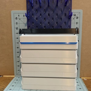 May include: A purple pegboard with vertical hooks sits behind a stack of five white rectangular storage bins. A black horizontal bar rests on the top bin. The unit is placed on a light green cutting mat with a grid marked in inches.