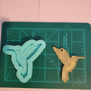 May include: A silicone mold of a hummingbird, with a gold-colored hummingbird on a green cutting mat with a grid pattern. The cutting mat has the text "Tech Tools CUTTING MAT GRID SIZE: 4"X6""