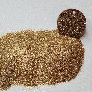 Op de afbeelding: Een hoop fijne gouden glitter met een iriserende glans. De glitter wordt getoond in een kleine hoop met een monster van de glitter in een ronde container.