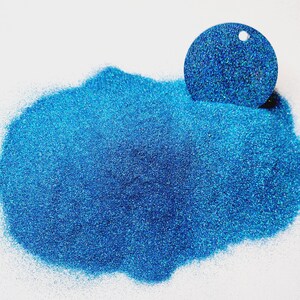 Puede incluir: Un montón de purpurina azul con brillo iridiscente. La purpurina está extendida sobre una superficie blanca. También se muestra una pequeña muestra redonda de la purpurina.