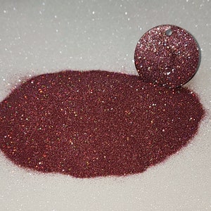 Puede incluir: Un montón de purpurina burdeos brillante con un dije circular a juego. La purpurina tiene una textura fina y refleja la luz, creando un efecto brillante. El dije tiene un pequeño agujero en la parte superior, lo que sugiere que podría usarse como colgante o adorno.