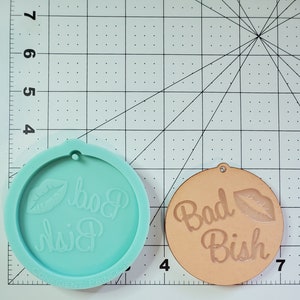 Op de afbeelding: Een lichtblauwe siliconen mal met de tekst "Bad Bish" en een rode lipafdruk. De mal ligt naast een lichtbruine houten cirkel met dezelfde tekst en lipafdruk.