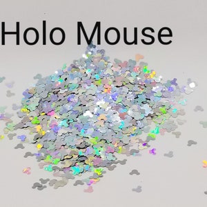 Puede incluir: Un montón de purpurina iridiscente con forma de cabezas de Mickey Mouse. La purpurina es plateada con un brillo arcoíris. El texto "Holo Mouse" está impreso en negro encima de la purpurina.