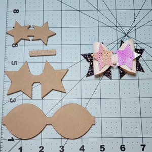 May include: A collection of craft supplies for making hair bows. The image shows various cut-out shapes in tan, including star shapes and bow outlines. A finished bow with glitter and iridescent accents is also present.