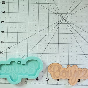 May include: A silicone mold and a finished resin ornament, both with the word "Boujee" in a cursive font. The mold is light blue and the ornament is brown.