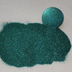 Puede incluir: Primer plano de un montón de purpurina verde azulado brillante junto a un dije circular verde azulado con un pequeño agujero en la parte superior. La purpurina está esparcida sobre una superficie blanca y brillante, creando un efecto brillante.