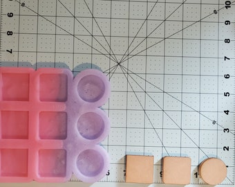May include: A pink and purple silicone mold with square and circular cavities, alongside three small, tan leather pieces. The mold is designed for crafting, with a grid background for measurement. The leather pieces are a square, a rounded square, and a circle.
