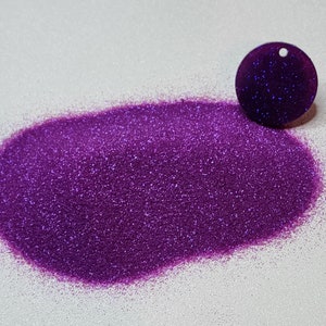 Puede incluir: Un montón de purpurina morada vibrante junto a un dije circular morado oscuro con un pequeño agujero en la parte superior. La purpurina tiene una calidad brillante y reflectante, y el dije parece estar hecho de un material liso.