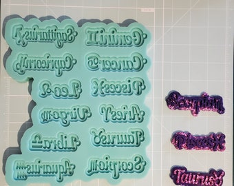 May include: A teal blue silicone mold with 12 zodiac signs, including Sagittarius, Gemini, Cancer, Leo, Virgo, Aries, Libra, Taurus, Scorpio, Pisces, Capricorn, and Aquarius.  The mold is next to 3 purple glitter zodiac signs: Scorpio, Pisces, and Taurus.