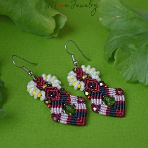 Op de afbeelding: Een paar handgemaakte macramé oorbellen. De oorbellen hebben een geometrisch ontwerp met rode, zwarte en witte draden, geaccentueerd met kleine kralen in verschillende kleuren. De oorbellen hebben een haaksluiting en staan tegen een groene achtergrond.