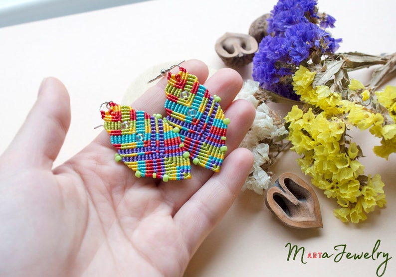 May include: A pair of colourful, geometrically patterned earrings made with macrame and beads. The earrings are in the shape of a teardrop and have a silver hook.