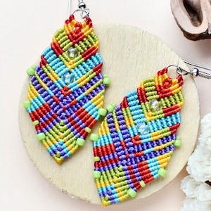 May include: A pair of colourful, geometrically patterned earrings made with macrame and beads. The earrings are in the shape of a teardrop and have a silver hook. The earrings are on a wooden surface with a white background. The text "PDF TUTORIAL" and "Maa Jewelry ART" are visible in the image.
