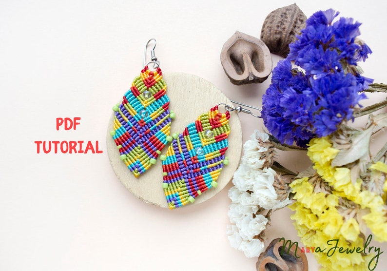 May include: A pair of colourful, geometrically patterned earrings made with macrame. The earrings are on a wooden surface with dried flowers in the background. The text "PDF TUTORIAL" is visible in the image.