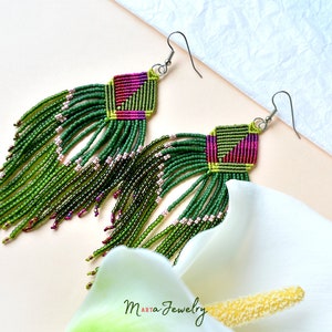 May include: Green and pink beaded earrings with a geometric pattern and fringe. The earrings are hanging from silver hooks.