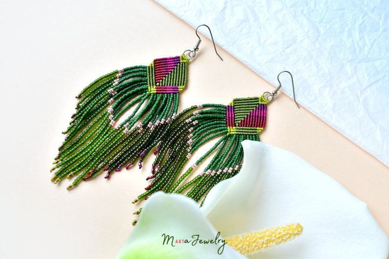 May include: Green and pink beaded earrings with a geometric pattern and fringe. The earrings are hanging from silver hooks.