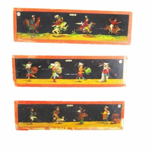 May include: Three vintage paper slides with colorful illustrations of people on horseback, marching in a parade, and working in a shop. The slides are in wooden frames with red and black borders.