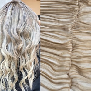 May include: A close-up of a weft of blonde human hair extensions. The hair is wavy and has a light blonde color with darker blonde highlights.