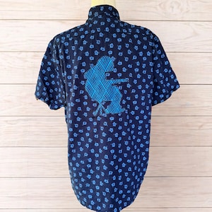 Könnte beinhalten: Marineblaues kurzärmeliges Hemd mit Blumenmuster und einer blauen Skifahrer-Grafik auf dem Rücken. Das Hemd wird auf einer Schaufensterpuppe präsentiert. Das Hemd ist aus einem weichen, bequemen Material gefertigt.