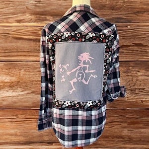 Op de afbeelding: Geruit flanellen overhemd in marineblauw, roze en wit, weergegeven op een mannequin. De achterkant heeft een grijze patch met een roze afbeelding en bloemmotieven. Het overhemd heeft lange mouwen en een knoopsluiting.