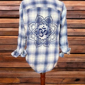 Puede incluir: Camisa abotonada a cuadros azules y blancos con un gran símbolo Om en la espalda. El símbolo Om es un diseño circular detallado en azul oscuro. La camisa se exhibe en un maniquí.