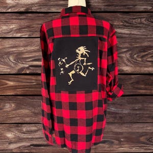 May include: Red and black plaid flannel shirt with a black square patch on the back featuring a gold graphic of a stylized figure. The shirt is displayed on a wooden mannequin.