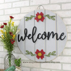 Pu&ograve; includere: Un cartello di benvenuto rotondo in legno con sfondo grigio e lettere nere. Il cartello &egrave; decorato con fiori rossi e gialli e foglie verdi. Il testo sul cartello dice "benvenuto".