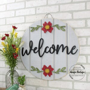 Pu&ograve; includere: Un cartello di benvenuto rotondo in legno con sfondo grigio e strisce bianche dipinte. La parola "benvenuto" &egrave; scritta in lettere corsive nere. Il cartello &egrave; decorato con fiori rossi e gialli e foglie verdi.