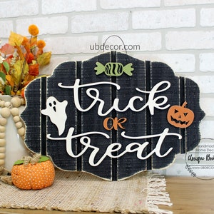 May include: Black wooden sign with the words "trick or treat" in white script. The sign is decorated with a white ghost, a green candy, and an orange pumpkin with a jack-o-lantern face. The sign is hanging on a white brick wall.