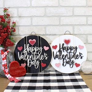 May include: Two round wooden signs with the text "Happy Valentine's Day" in black script. The signs are decorated with pink and red hearts. One sign has a black background and the other has a white background.