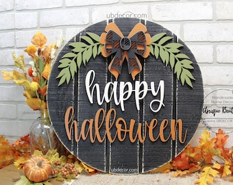 Op de afbeelding: Een rond houten bord met de woorden "happy halloween" in witte en oranje letters. Het bord is versierd met een zwart-witte geruite strik met oranje accenten en groene bladeren.
