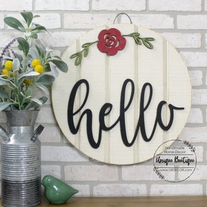 May include: A round wooden sign with a white background and a black cursive "hello" on it. The sign is decorated with a red paper rose and green leaves. The sign is hanging on a brick wall.