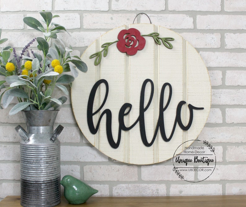 May include: A round wooden sign with a white background and a black cursive "hello" on it. The sign is decorated with a red paper rose and green leaves. The sign is hanging on a brick wall.