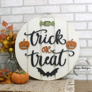 May include: A round wooden sign with the words "Trick or Treat" in black script. The sign is decorated with orange and green pumpkins, a bat, and a candy. The sign is on a white brick wall.