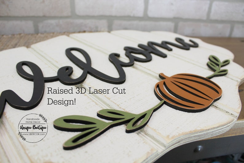 May include: A white wooden sign with the word "Welcome" in black raised 3D letters. The sign also features a brown pumpkin and green leaves, all laser cut. The text "Raised 3D Laser Cut Design!" is printed below the welcome sign. The sign is handmade by Unique Boutique.