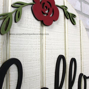 May include: White wooden round sign with the word "hello" in black lettering. The sign is decorated with a red rose and green leaves.
