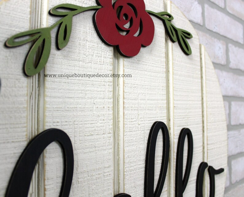 May include: White wooden round sign with the word "hello" in black lettering. The sign is decorated with a red rose and green leaves.