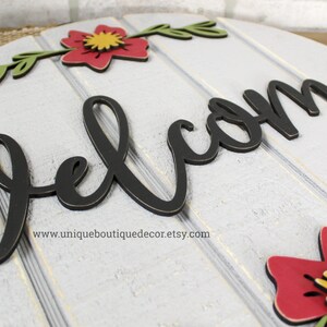 Pu&ograve; includere: Un cartello rotondo in legno bianco con la parola "welcome" in lettere nere in stile corsivo. Il cartello &egrave; decorato con fiori rossi e verdi.