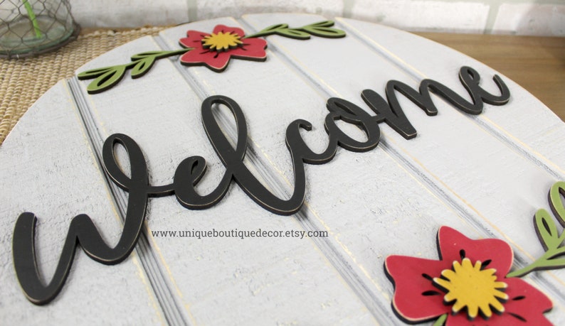 Pu&ograve; includere: Un cartello rotondo in legno bianco con la parola "welcome" in lettere nere in stile corsivo. Il cartello &egrave; decorato con fiori rossi e verdi.
