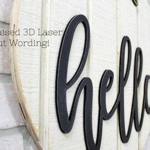 May include: A white wooden round sign with the word "hello" in black raised 3D laser cut lettering. The sign is mounted on a white brick wall. The text "Raised 3D Laser Cut Wording!" is visible in the top left corner of the image.