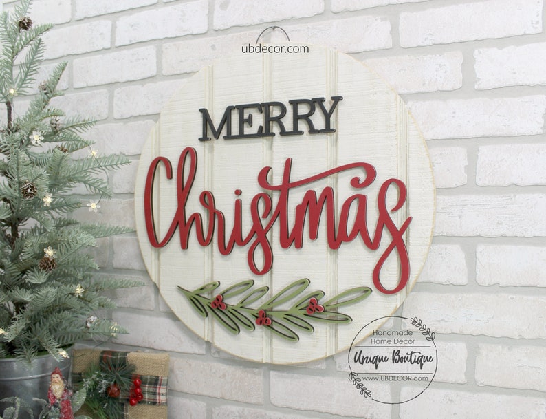 K&ouml;nnte beinhalten: Ein rundes wei&szlig;es Holzschild mit den Worten "Merry Christmas" in schwarzer und roter Schrift. Das Schild hat unten einen gr&uuml;nen und roten Stechpalmenzweig mit Beeren.