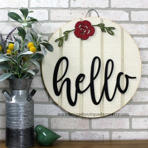May include: A round wooden sign with a white background and black vertical stripes. The word "hello" is written in black cursive script. A red paper flower with green leaves is attached to the top of the sign.