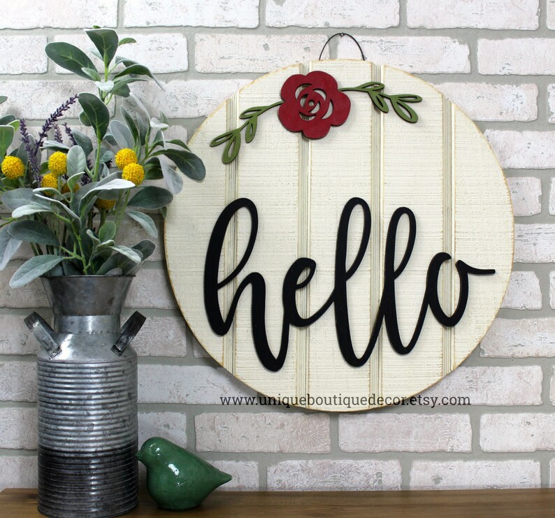 May include: A round wooden sign with a white background and black vertical stripes. The word "hello" is written in black cursive script. A red paper flower with green leaves is attached to the top of the sign.