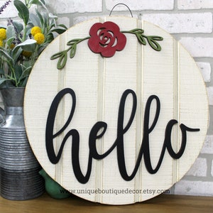 May include: A round wooden sign with a white background and black script lettering that says "hello". A red paper flower with green leaves is attached to the top of the sign.