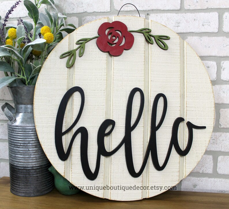 May include: A round wooden sign with a white background and black script lettering that says "hello". A red paper flower with green leaves is attached to the top of the sign.