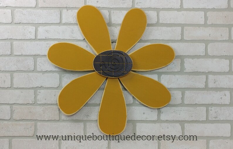 Puede incluir: Una gran margarita amarilla mostaza con un centro blanco y negro. La margarita est&aacute; hecha de madera y tiene un estilo r&uacute;stico, de granja.