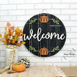 May include: A round wooden welcome sign with a black background and white lettering. The sign is decorated with fall-themed elements, including pumpkins and leaves. The sign is hanging on a white brick wall. The text on the sign reads "welcome".
