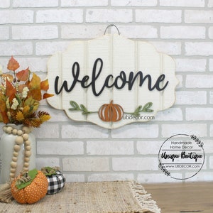 May include: A white wooden welcome sign with black script lettering and a carved pumpkin. The sign is hanging on a white brick wall. The sign is decorated with green leaves and a brown pumpkin. The sign is from Unique Boutique, a handmade home decor shop.