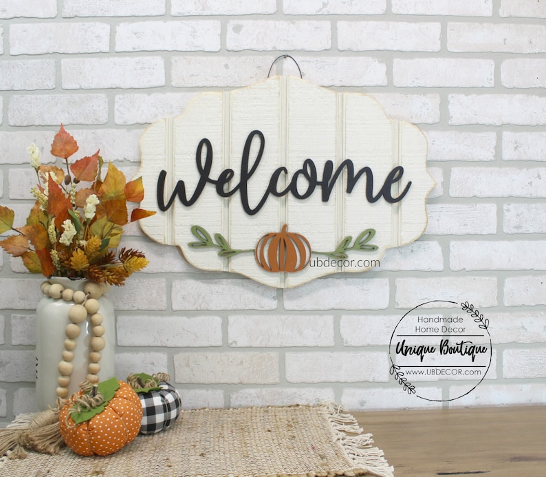 May include: A white wooden welcome sign with black script lettering and a carved pumpkin. The sign is hanging on a white brick wall. The sign is decorated with green leaves and a brown pumpkin. The sign is from Unique Boutique, a handmade home decor shop.