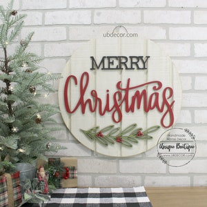 K&ouml;nnte beinhalten: Ein rundes Holzschild mit den Worten "Merry Christmas" in roten und schwarzen Buchstaben. Das Schild ist mit gr&uuml;nen Bl&auml;ttern und roten Beeren verziert. Das Schild befindet sich an einer wei&szlig;en Backsteinwand mit einem kleinen Weihnachtsbaum im Vordergrund.