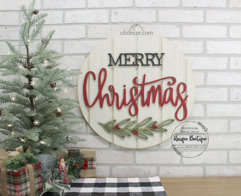 K&ouml;nnte beinhalten: Ein rundes Holzschild mit den Worten "Merry Christmas" in roten und schwarzen Buchstaben. Das Schild ist mit gr&uuml;nen Bl&auml;ttern und roten Beeren verziert. Das Schild befindet sich an einer wei&szlig;en Backsteinwand mit einem kleinen Weihnachtsbaum im Vordergrund.
